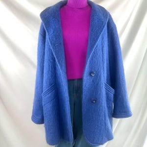 May include: A blue coat with a wide collar and two buttons. The coat has two front pockets and is paired with a purple turtleneck sweater and blue jeans. The coat is made of a fuzzy material.