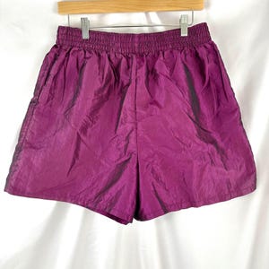 May include: Plum-colored athletic shorts with an elastic waistband. The shorts are made of a shiny, crinkled fabric and hang from a wooden hanger. The shorts have a relaxed fit and are suitable for various activities.