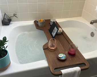 May include: A dark wood bathtub tray with a handle, holding a mug, a small bowl, a candle, a glass of wine, and an e-reader. A white towel hangs from the tray. The bathtub is filled with water and bubbles.