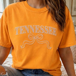 May include: A bright orange short-sleeved t-shirt featuring the word TENNESSEE embroidered in white thread across the chest, positioned directly above a white embroidered graphic of a decorative ribbon bow with flowing ends.