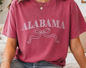 Peut inclure: Un t-shirt à manches courtes et col rond d'une couleur rouge cramoisi délavé. Le devant du t-shirt présente le mot ALABAMA brodé en lettres capitales blanches, placé au-dessus d'un motif linéaire blanc représentant un nœud décoratif.