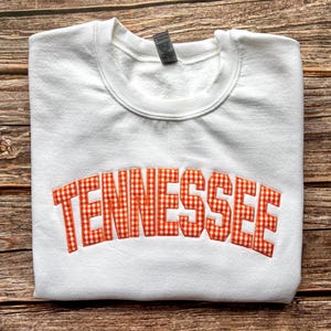 May include: A white crewneck sweatshirt featuring the word TENNESSEE embroidered in an arched design across the chest. The lettering is filled with an orange and white gingham check pattern and outlined with a thick orange stitched border.