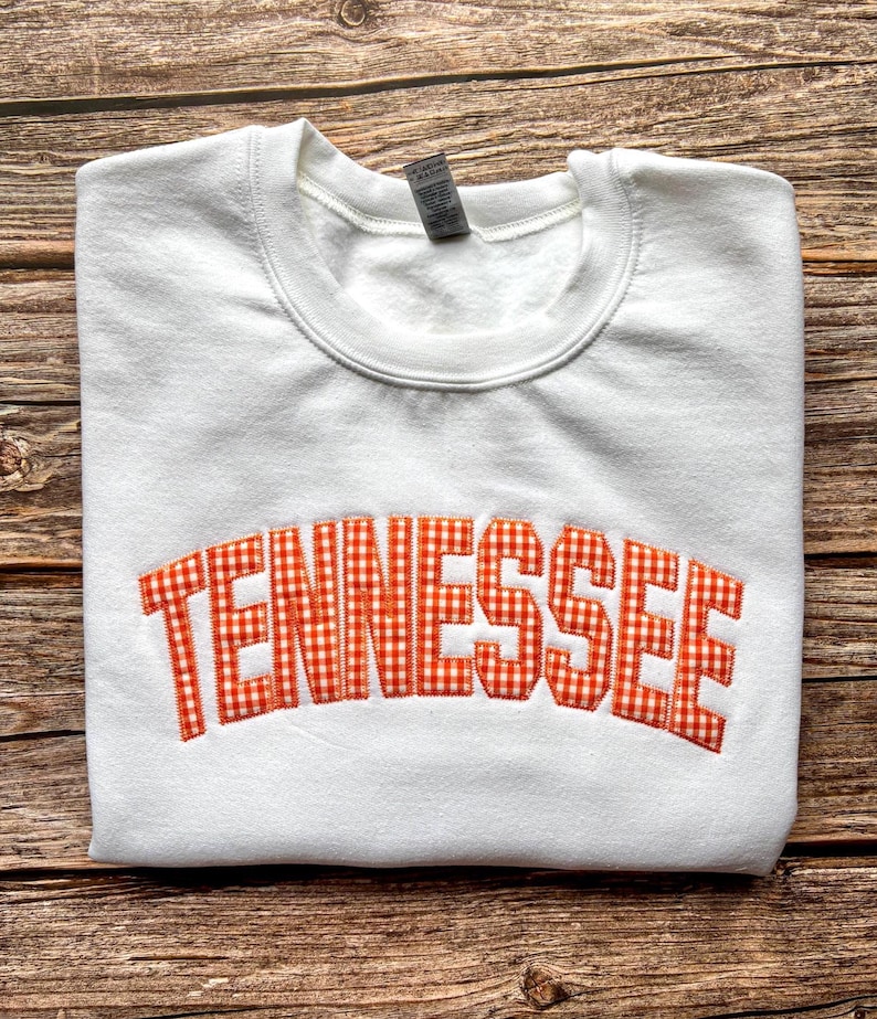 May include: A white crewneck sweatshirt featuring the word TENNESSEE embroidered in an arched design across the chest. The lettering is filled with an orange and white gingham check pattern and outlined with a thick orange stitched border.