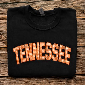 May include: A black crewneck sweatshirt featuring the word TENNESSEE in an arched layout across the chest. The letters are filled with an orange and white gingham check pattern and are outlined with a thick orange border stitched onto the fabric.