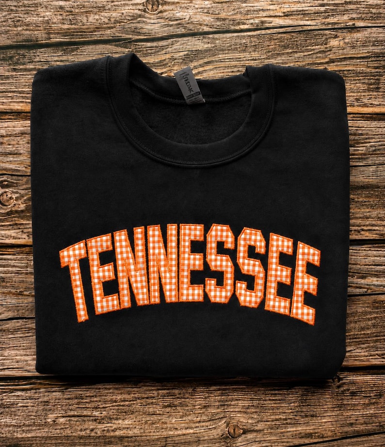 May include: A black crewneck sweatshirt featuring the word TENNESSEE in an arched layout across the chest. The letters are filled with an orange and white gingham check pattern and are outlined with a thick orange border stitched onto the fabric.