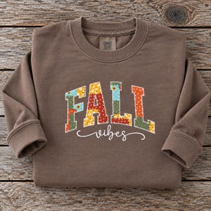 Könnte beinhalten: Ein braunes Langarm-Sweatshirt mit Rundhalsausschnitt, das das Wort FALL in großen, blockartigen, mehrfarbigen Buchstaben mit Blumen- und Punktemuster zeigt, wobei das Wort vibes in weißer Schreibschrift darunter steht.