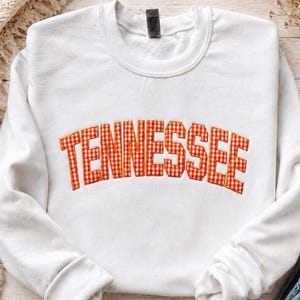 May include: A white crewneck sweatshirt featuring the word TENNESSEE arched across the chest in an orange and white gingham patterned fabric with a stitched border, showcasing a classic long-sleeve design with a ribbed collar and cuffs.