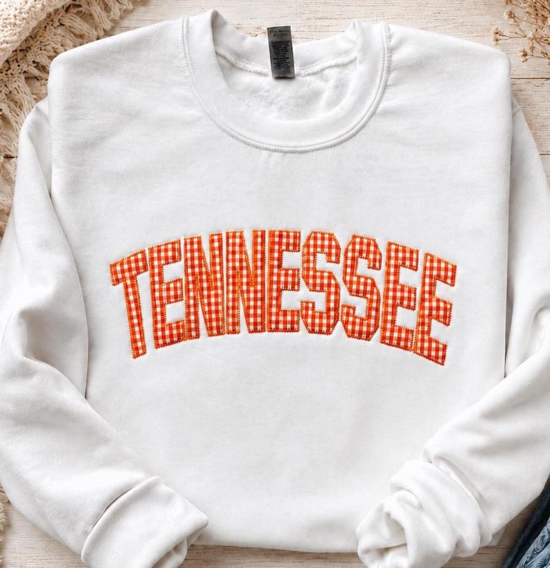 May include: A white crewneck sweatshirt featuring the word TENNESSEE arched across the chest in an orange and white gingham patterned fabric with a stitched border, showcasing a classic long-sleeve design with a ribbed collar and cuffs.