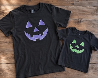 May include: Two black short-sleeved t-shirts featuring embroidered jack-o'-lantern faces. The larger shirt displays a purple pumpkin face, while the smaller shirt features a bright green pumpkin face, both centred on the chest area.