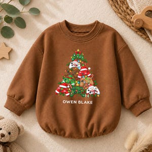 Op de afbeelding: Een bruin sweatshirt met lange mouwen en een afbeelding van een kerstboom opgebouwd uit diverse kleurrijke videogamecontrollers, ornamenten en lichtjes. De tekst OWEN BLAKE staat in witte hoofdletters onder de afbeelding gedrukt.