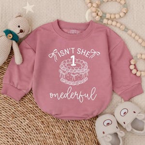Può includere: Un body da bambino color rosa polveroso con maniche lunghe. La parte anteriore presenta una grafica bianca di una torta con il numero 1 e la scritta "Isn't she 1 onederful". Un orsacchiotto e scarpine per bambini sono sullo sfondo.