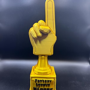 Peut inclure: Trophée jaune imprimé en 3D en forme de main avec l'index pointé vers le haut. La base du trophée porte l'inscription "Fantasy League Winner" en noir. Le trophée est posé sur un fond noir.