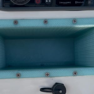 May include: A rectangular, light blue storage compartment with a textured surface, secured with silver screws. The compartment is recessed into a white surface, with a black cap and a stereo system visible above.