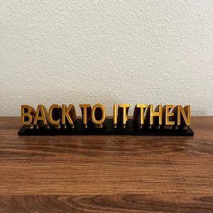 Può includere: Un cartello dorato stampato in 3D con la scritta "BACK TO IT THEN" in grassetto e maiuscolo. Le lettere sono montate su una base nera e il cartello è esposto su una superficie di legno.