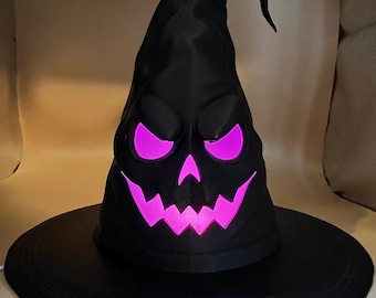 May include: A black witch hat with a pointed, slightly drooping top and a wide brim. The front of the hat features a carved, menacing face with sharp, jagged eyes and a wide, toothy grin that glows with a bright, vibrant pink light from within.