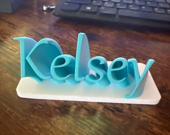 May include: A 3D-printed nameplate with the name "Kelsey" in light blue letters. The letters are raised and sit on a white rectangular base. The nameplate is on a dark wooden surface.