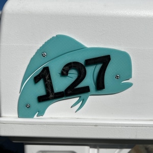 May include: A turquoise fish-shaped house number sign with the number "127" in black. The sign is mounted on a white mailbox. The fish has a textured pattern and is secured with silver screws.