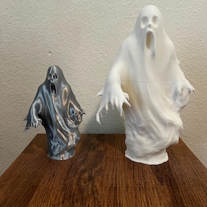 May include: Two ghost sculptures, one metallic silver and the other white, stand on a wooden surface. The ghosts have open mouths and outstretched arms, with a flowing, draped appearance. The larger ghost is approximately 12 inches tall.