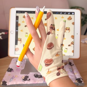 Peut inclure: Un gant de dessin beige avec un motif de raton laveur et d'aliments de dessin animé est porté tout en tenant un stylet jaune. Le stylet est près d'une tablette affichant une illustration numérique. La tablette repose sur un coussin rose avec des illustrations de chats.