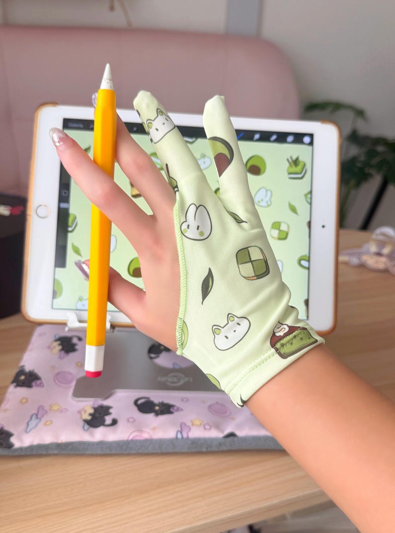 May include: A light green drawing glove with cartoon avocado, bunny, and cake designs. A yellow pencil with a white tip is held near a tablet displaying a digital drawing. The tablet rests on a pink pad with cat illustrations.