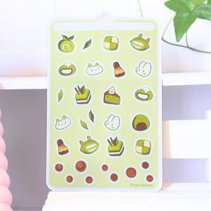 Puede incluir: Una hoja de pegatinas rectangular con fondo verde claro que presenta varias ilustraciones de postres de matcha, incluyendo pasteles, mochi y caras de animales, junto con perlas de boba marrones y el texto POLYMERNAI.