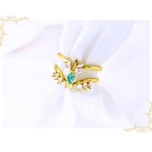 May include: A gold-toned ring with a double band design. The ring features a central teal-colored gemstone and is adorned with white, marquise-cut stones. The ring is set against a white background.