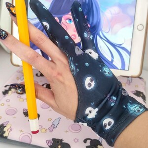 May include: A dark blue artist's glove with a pattern of white ghost and butterfly illustrations. A yellow stylus with a white tip is held in the glove. The glove is designed for digital art and drawing on a tablet.