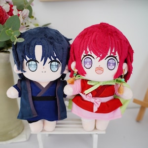 May include: Two plush dolls, one with dark blue hair and clothing, the other with red hair and a red and pink outfit. Both dolls have large, light blue eyes and are sitting on a white bench. The dolls are anime-style.