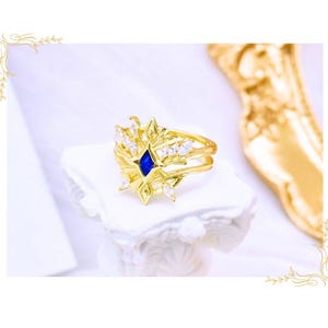 May include: A gold-colored ring with a starburst design, featuring a central blue gemstone and surrounding clear crystals. The ring is displayed on a white, ornate pedestal, showcasing its intricate details and craftsmanship.