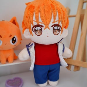 May include: A plush doll with orange hair, wearing a red shirt, blue shorts, and a white jacket. The doll has large, expressive eyes and is standing next to a smaller orange plush figure.