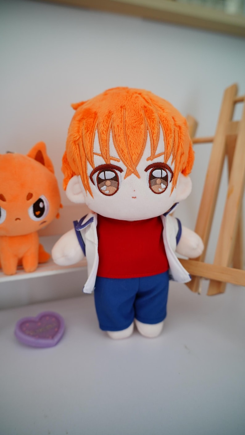 May include: A plush doll with orange hair, wearing a red shirt, blue shorts, and a white jacket. The doll has large, expressive eyes and is standing next to a smaller orange plush figure.