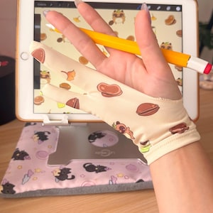 May include: A beige digital artist's glove with a pattern of brown food and cartoon characters is held over a tablet with a stylus. The tablet is on a stand with a pink and gray cat-themed cover. A yellow stylus with a red tip rests on the hand.