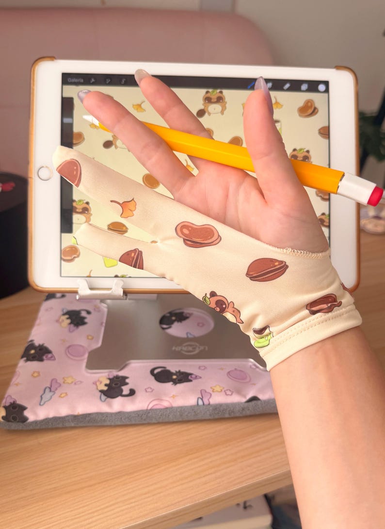 May include: A beige digital artist's glove with a pattern of brown food and cartoon characters is held over a tablet with a stylus. The tablet is on a stand with a pink and gray cat-themed cover. A yellow stylus with a red tip rests on the hand.