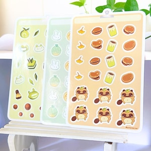 Puede incluir: Tres hojas de pegatinas rectangulares con diversas ilustraciones. La hoja izquierda tiene iconos verdes de frutas y pasteles. La hoja central muestra caras de conejo blancas y patrones de melón. La hoja derecha muestra hojas de ginkgo, dorayaki y personajes tanuki de color tostado con el texto @polymernai.
