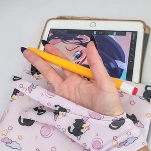 May include: A light pink tablet sleeve featuring a repeating pattern of black cats, stars, clouds, and moons. A yellow stylus rests on the sleeve, near a tablet displaying digital art. The sleeve is designed to protect the tablet.