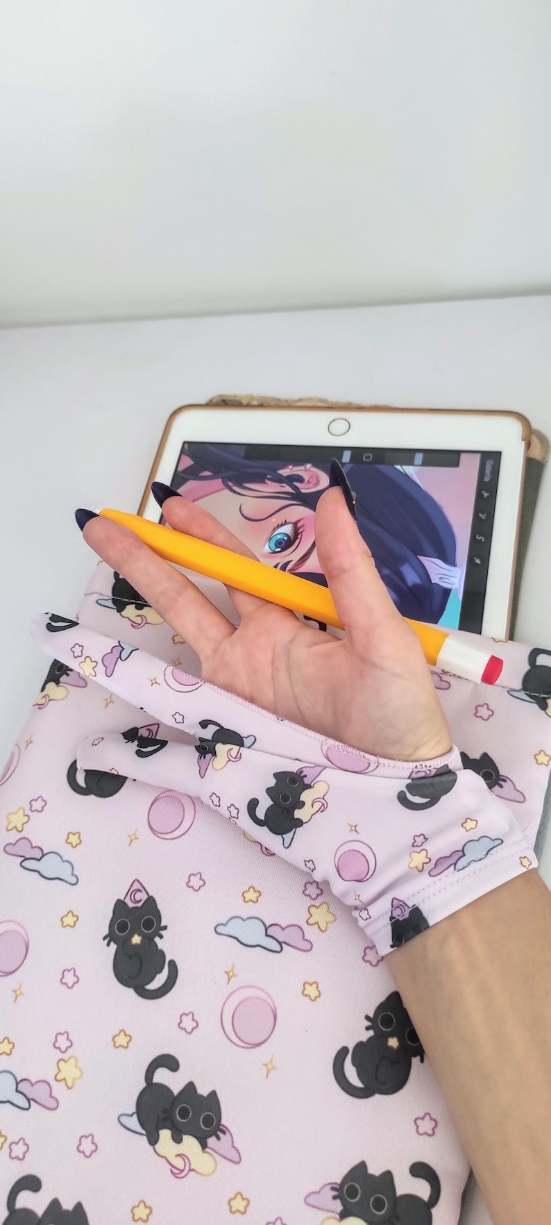 May include: A light pink tablet sleeve featuring a repeating pattern of black cats, stars, clouds, and moons. A yellow stylus rests on the sleeve, near a tablet displaying digital art. The sleeve is designed to protect the tablet.