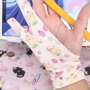 May include: A light pink artist's glove with a pattern of yellow ducklings, strawberries, and flowers. A yellow stylus is held in the hand, with a digital drawing tablet in the background. The glove is designed for digital art.