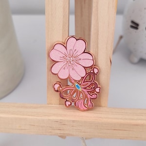 Puede incluir: Un pin de esmalte rosa con una flor de cerezo y un lazo decorativo. El pin tiene un contorno dorado y una pequeña gema azul en el centro del lazo. La flor de cerezo tiene un acabado brillante.