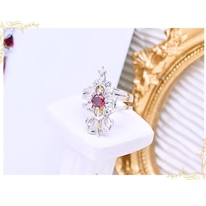 May include: A silver ring with an ornate design, featuring a central red gemstone. The ring is displayed on a white pedestal, with a gold-colored decorative frame in the background. The ring's design includes intricate filigree and a crown-like top.