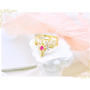 May include: A gold-colored ring with a central, teardrop-shaped red gemstone. The ring features a floral design with clear, white gemstones and is displayed on a white, decorative pedestal. The background includes soft pink and white elements.