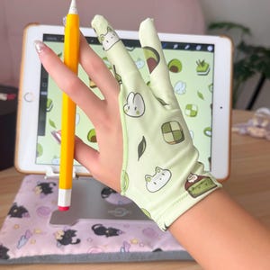 Może przedstawiać: Jasnozielona rękawica do rysowania z kreskówkowymi wzorami awokado, królika i ciasta. Żółty ołówek z białą końcówką trzymany jest w pobliżu tabletu wyświetlającego cyfrowy projekt artystyczny. Tablet spoczywa na różowej podkładce.