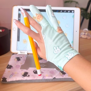 May include: A light blue drawing glove with bunny and bread graphics is worn while using a yellow stylus on a tablet. The tablet is in a pink case with black cat graphics. The background includes a wooden desk.