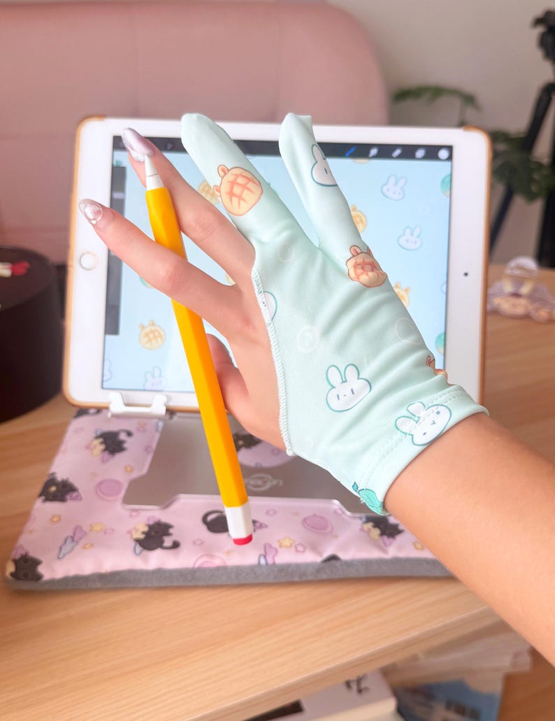May include: A light blue drawing glove with bunny and bread graphics is worn while using a yellow stylus on a tablet. The tablet is in a pink case with black cat graphics. The background includes a wooden desk.