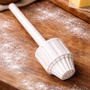 Puede incluir: Un prensador de pastelería blanco con un mango largo y una base acanalada en forma de magdalena descansa sobre una tabla de cortar de madera espolvoreada con harina. Ingredientes como huevos, mantequilla y harina sugieren hornear.