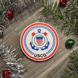 Może przedstawiać: Okrągła naszywka z emblematem USCG, z czerwoną i białą obwódką, niebieskimi kotwicami i napisem "USCG". Naszywka otoczona jest dekoracjami świątecznymi, w tym tinsel, dzwonkami i gałązkami sosny.