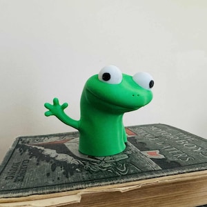 May include: A green frog-shaped bookmark with large white eyes and a raised arm. The bookmark is placed on top of a vintage book with the title "The Motor Boys Strange Waters" visible.