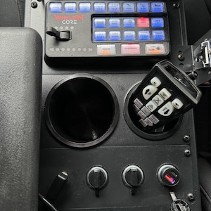 以下が含まれることがあります： 「Whelen Core」の文字と、「Wail」、「Yelp」、「Horn」などの機能が記載された複数のボタンが付いた黒いコントロールパネル。カップホルダー、マイク、さまざまなスイッチとダイヤルが含まれています。