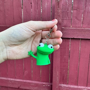 May include: A bright green frog keychain with a silver clasp. The frog has large white eyes with black pupils and is waving one arm. The background is a red wooden fence.