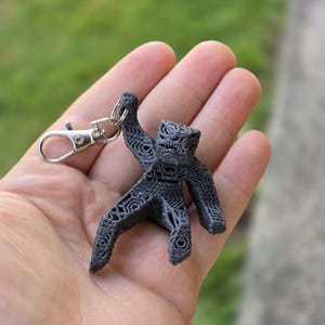 May include: A black 3D-printed keychain featuring a stylized figure with intricate geometric patterns. The figure is attached to a silver-colored metal clasp. The background is a blurred view of green foliage.