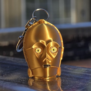 May include: A metallic gold-colored keychain shaped like the head of a humanoid robot. The figure features a smooth, rounded dome, prominent circular eye sockets with yellow and black detailing, and a small rectangular mouth area. A silver metal ring is attached.